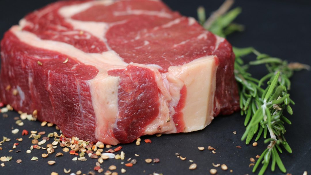 a steak ready for grilling next to seasoning and a sprig of rosemary